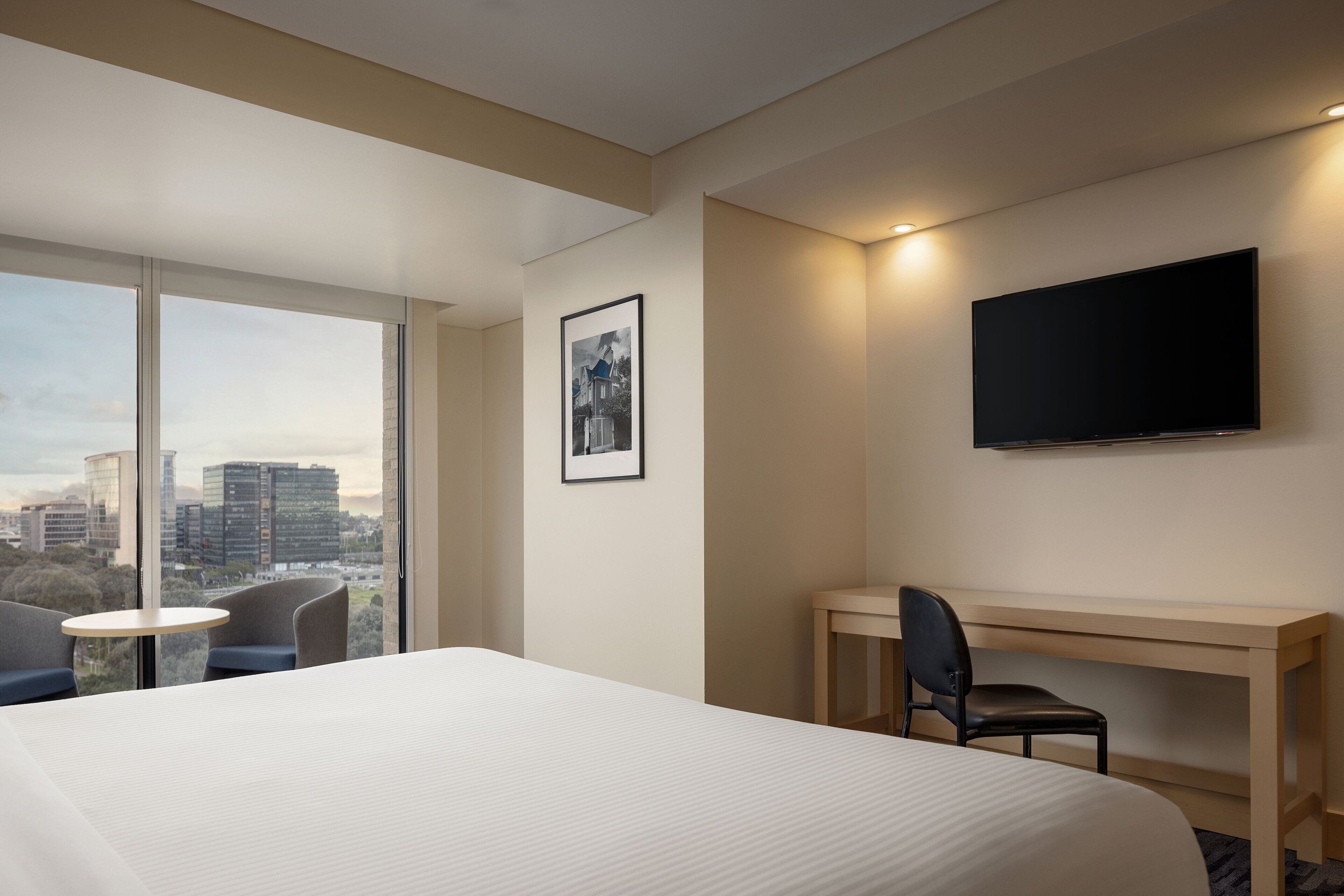 A hotel room with a large bed, a desk, a chair, a TV, and a window with a city view.