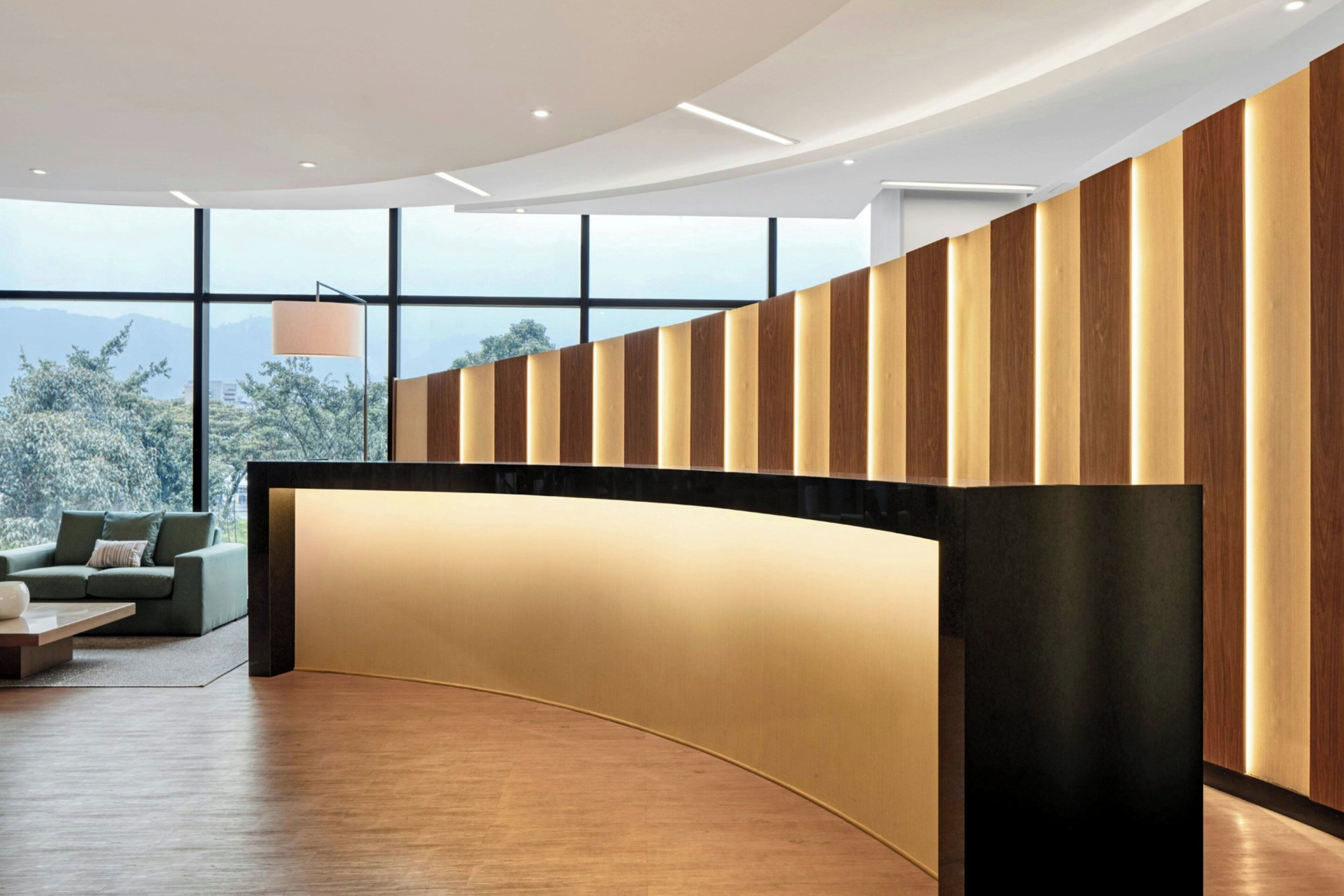 Un área de recepción moderna con un mostrador curvo, ventanas amplias que ofrecen vistas a árboles y montañas, y un sofá con una mesita de centro.