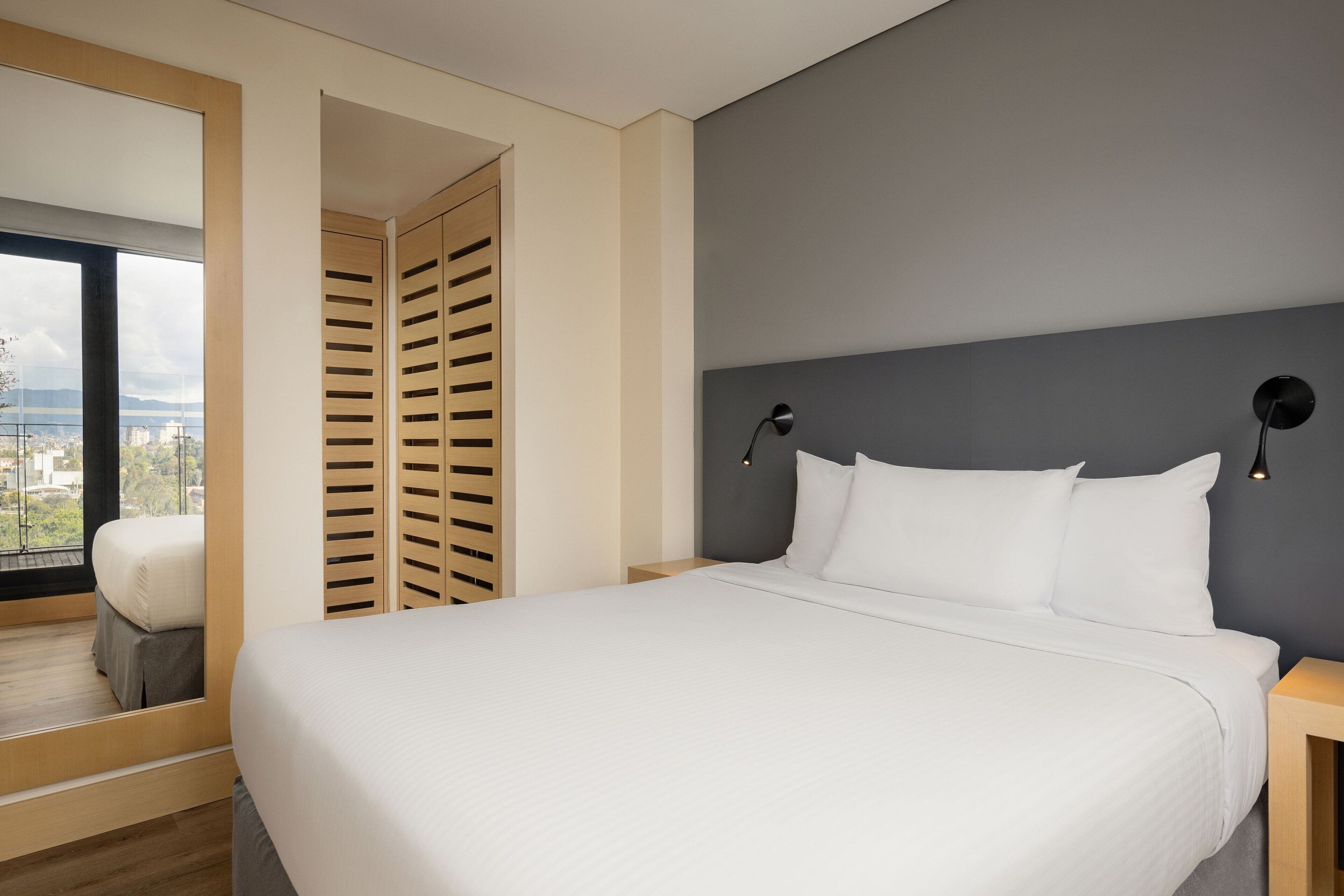 A modern hotel room with a large bed, a view through a window, and a wooden wardrobe.