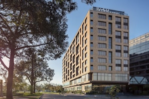 A modern hotel building with a brick facade and large windows, located in a landscaped area with trees and a clear sky.