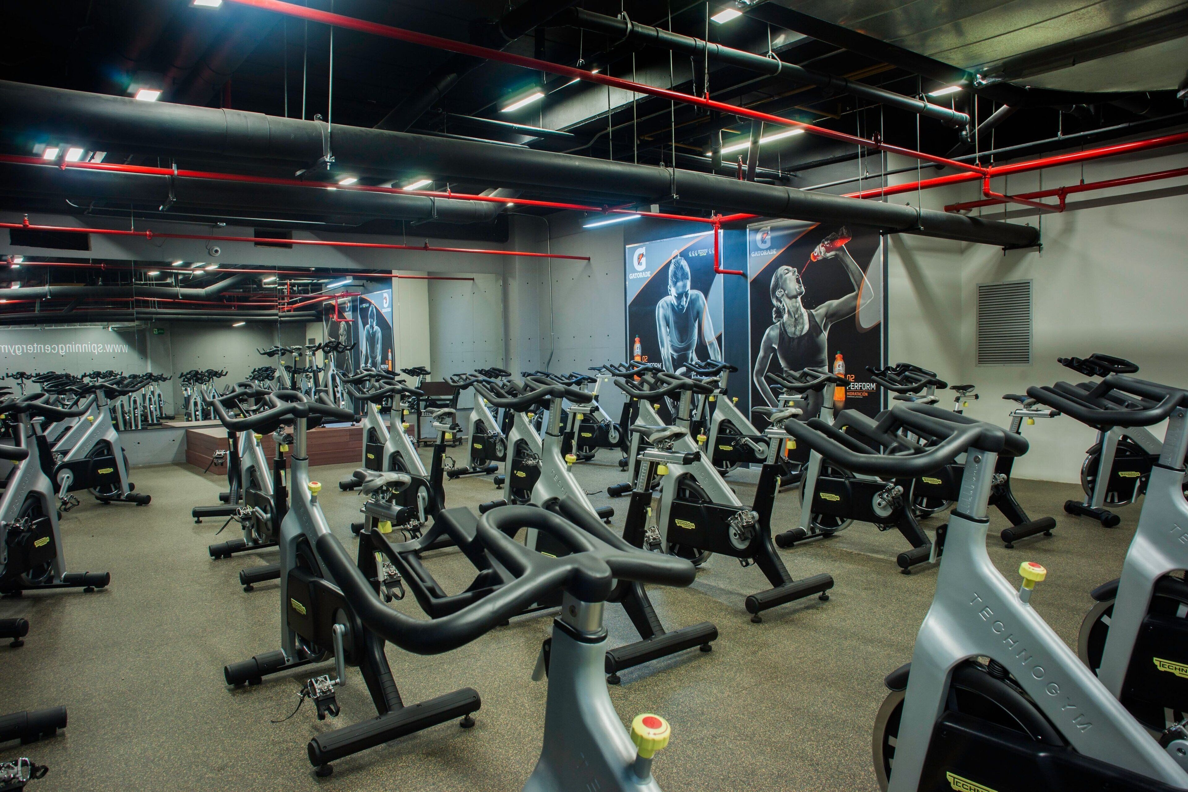 Un gimnasio con varias bicicletas estáticas y equipo de ejercicio, tuberías rojas en el techo y un cartel grande en la pared.