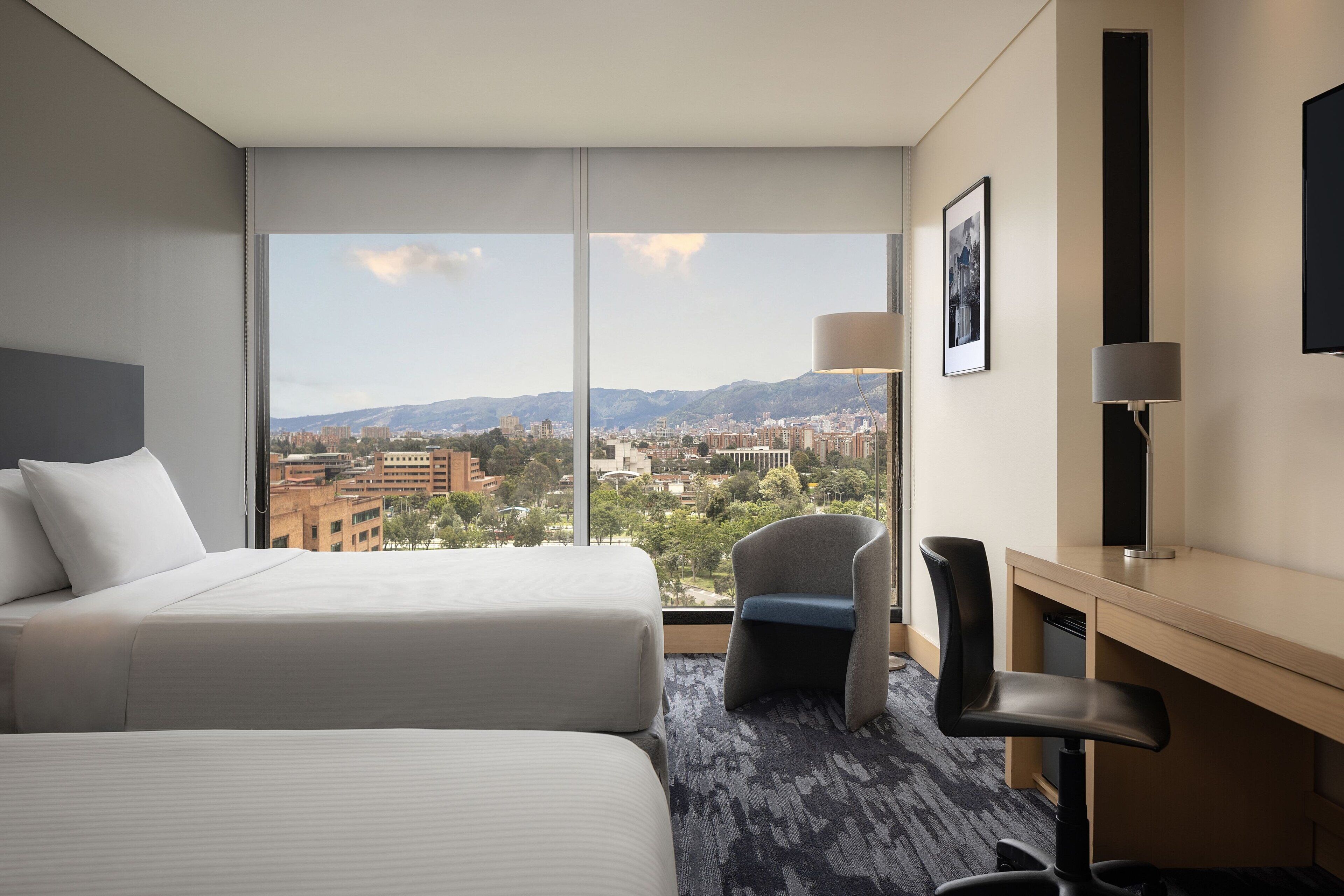 Quarto de hotel com duas camas, uma escrivaninha, uma cadeira, uma televisão e uma janela grande com vista para a cidade e montanhas.