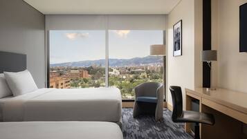 Quarto de hotel com duas camas, uma escrivaninha, uma cadeira, uma televisão e uma janela grande com vista para a cidade e montanhas.