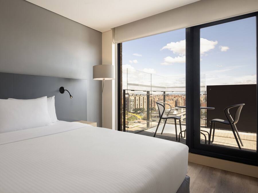 Una camera d'albergo moderna con un letto grande, un comodino, una lampada e un balcone con vista sulla città.