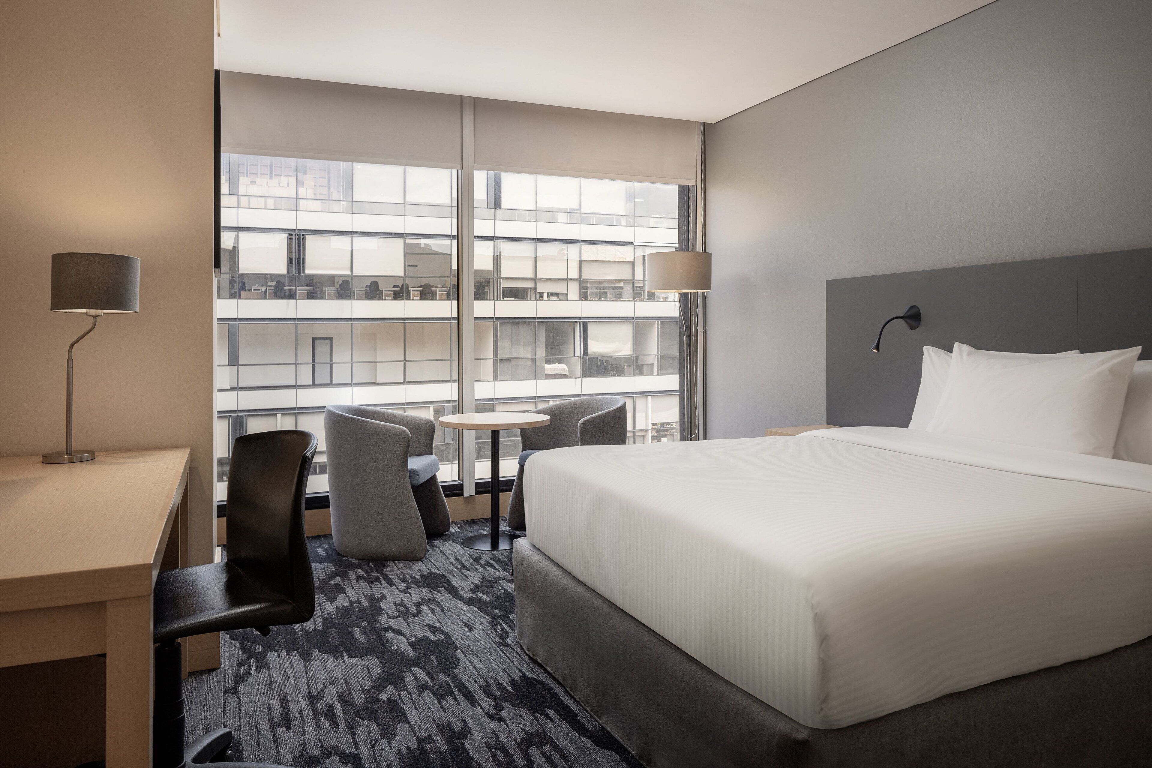 Hotelkamer met een groot bed, een bureau, twee stoelen, een kleine ronde tafel en uitzicht op een gebouw door grote ramen.