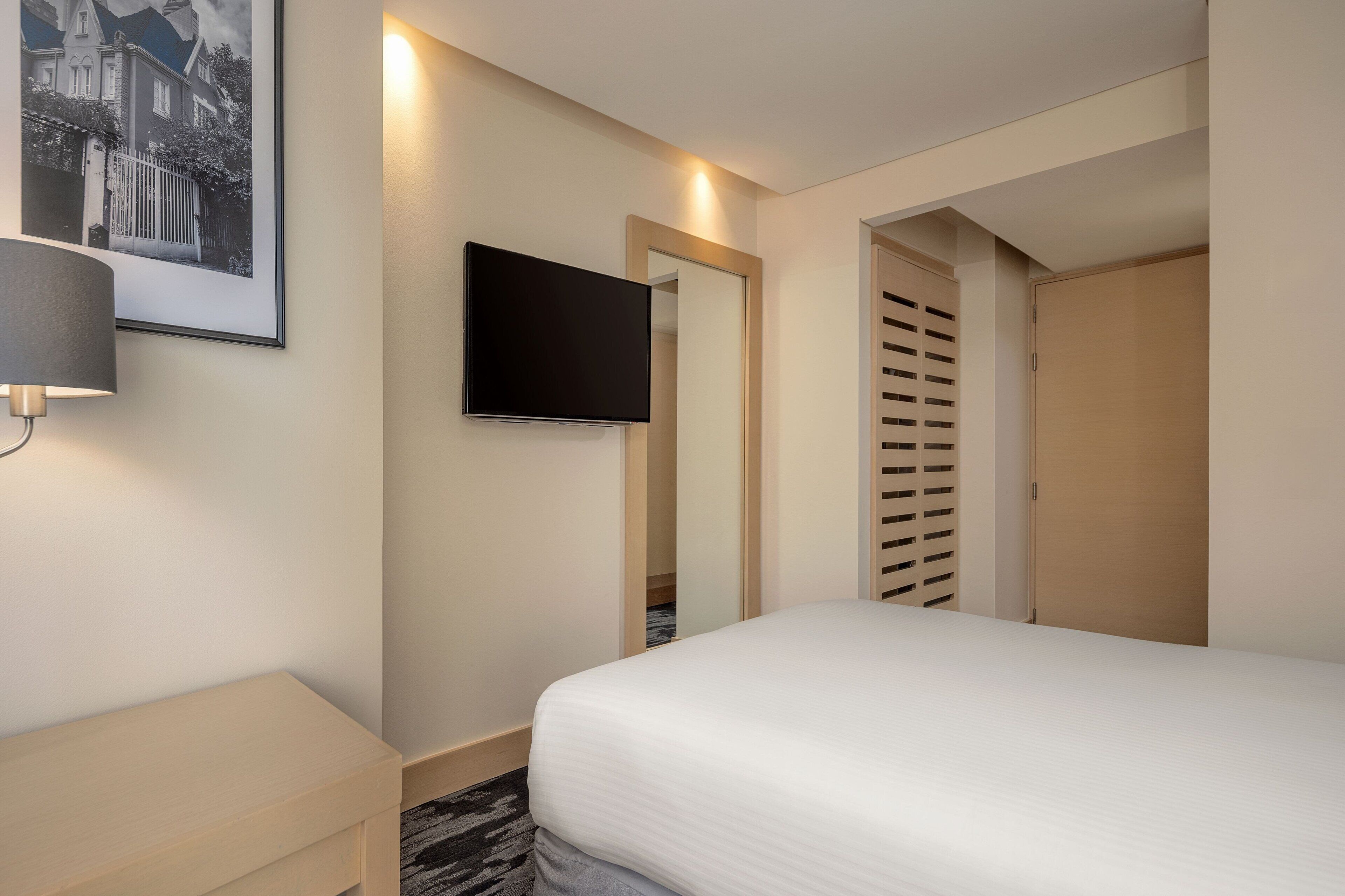 A hotel room with a bed, a bedside table, a lamp, a TV mounted on the wall, and a door with a louvered design.