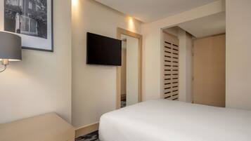 A hotel room with a bed, a bedside table, a lamp, a TV mounted on the wall, and a door with a louvered design.
