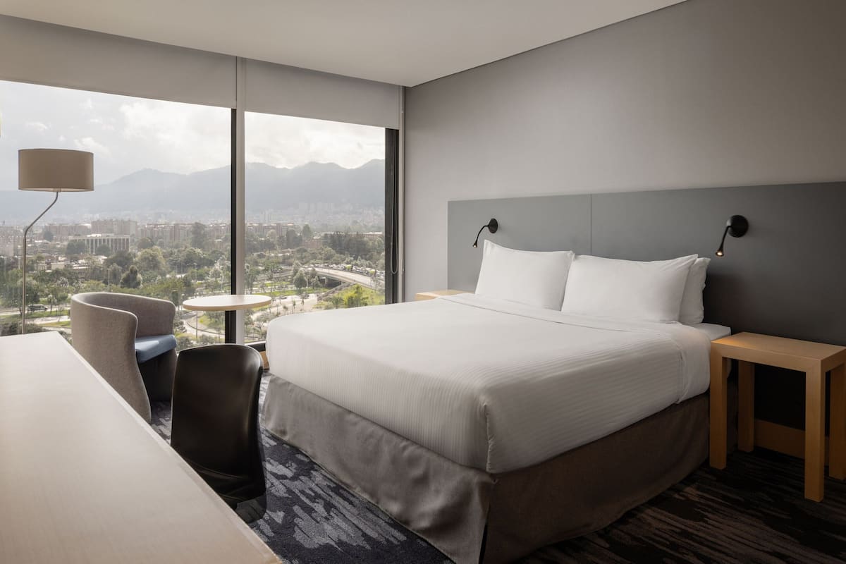A modern hotel room with a large bed, a desk, and a chair. There is a view of a cityscape through large windows.