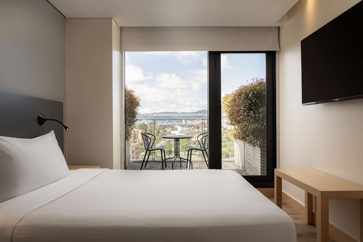 A modern hotel room with a large bed, a TV mounted on the wall, and a balcony with a view of the city and mountains.