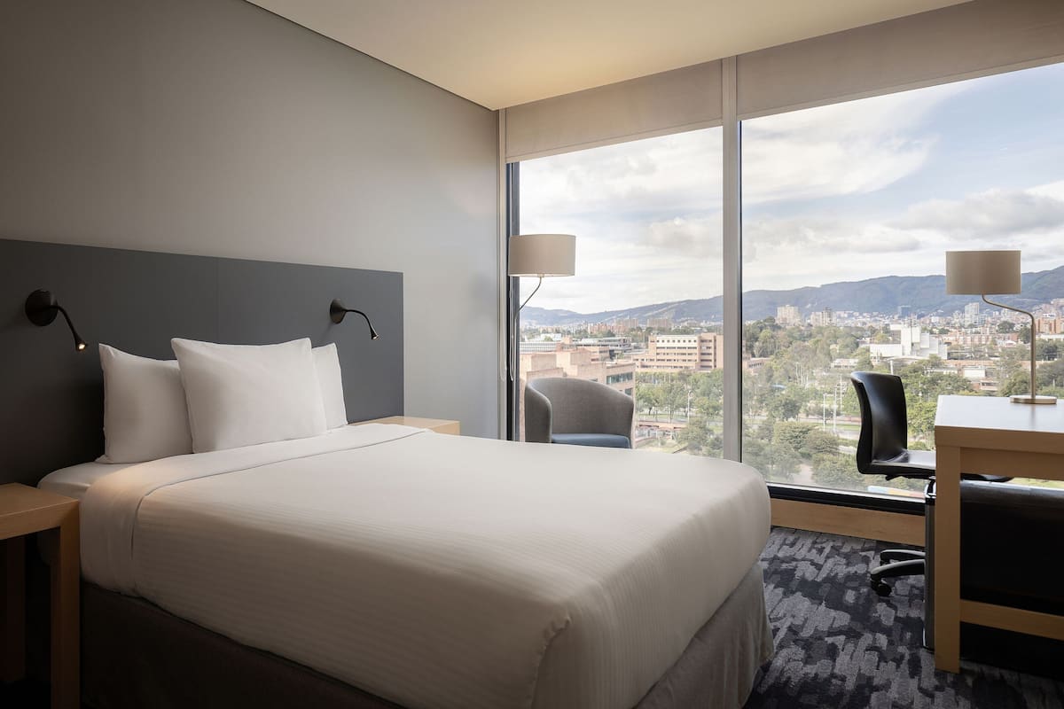 A hotel room with a large bed, two bedside lamps, a desk with a chair, and a view of a cityscape through large windows.