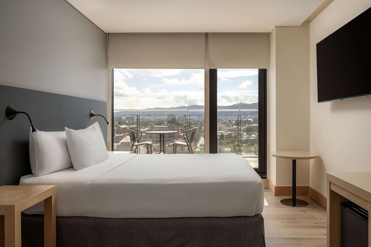 A modern hotel room with a large bed, a TV, a small table, and a balcony with a view of the city.