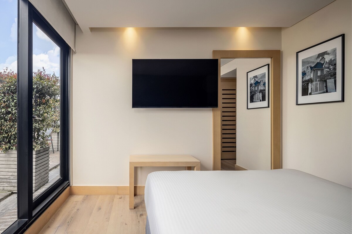 Un dormitorio con una cama, un televisor montado en la pared, un pequeño banco de madera y cuadros enmarcados.