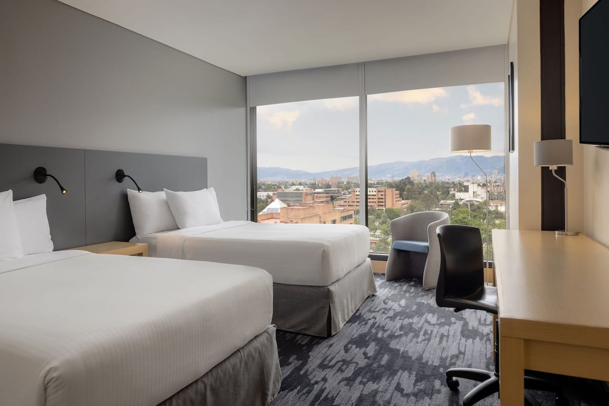 A hotel room with two beds, a desk, a chair, and a large window offering a city view.