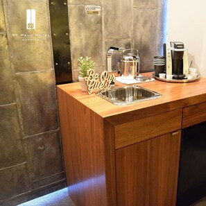 Cozinha compacta de hotel com uma pia de aço inoxidável, uma cafeteira e uma placa com a mensagem 'Seja nosso convidado'.