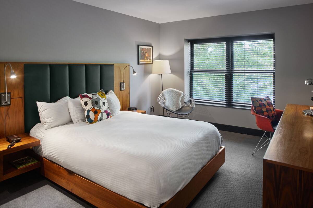 A modern hotel room with a large bed, a desk, a chair, and a window with blinds.