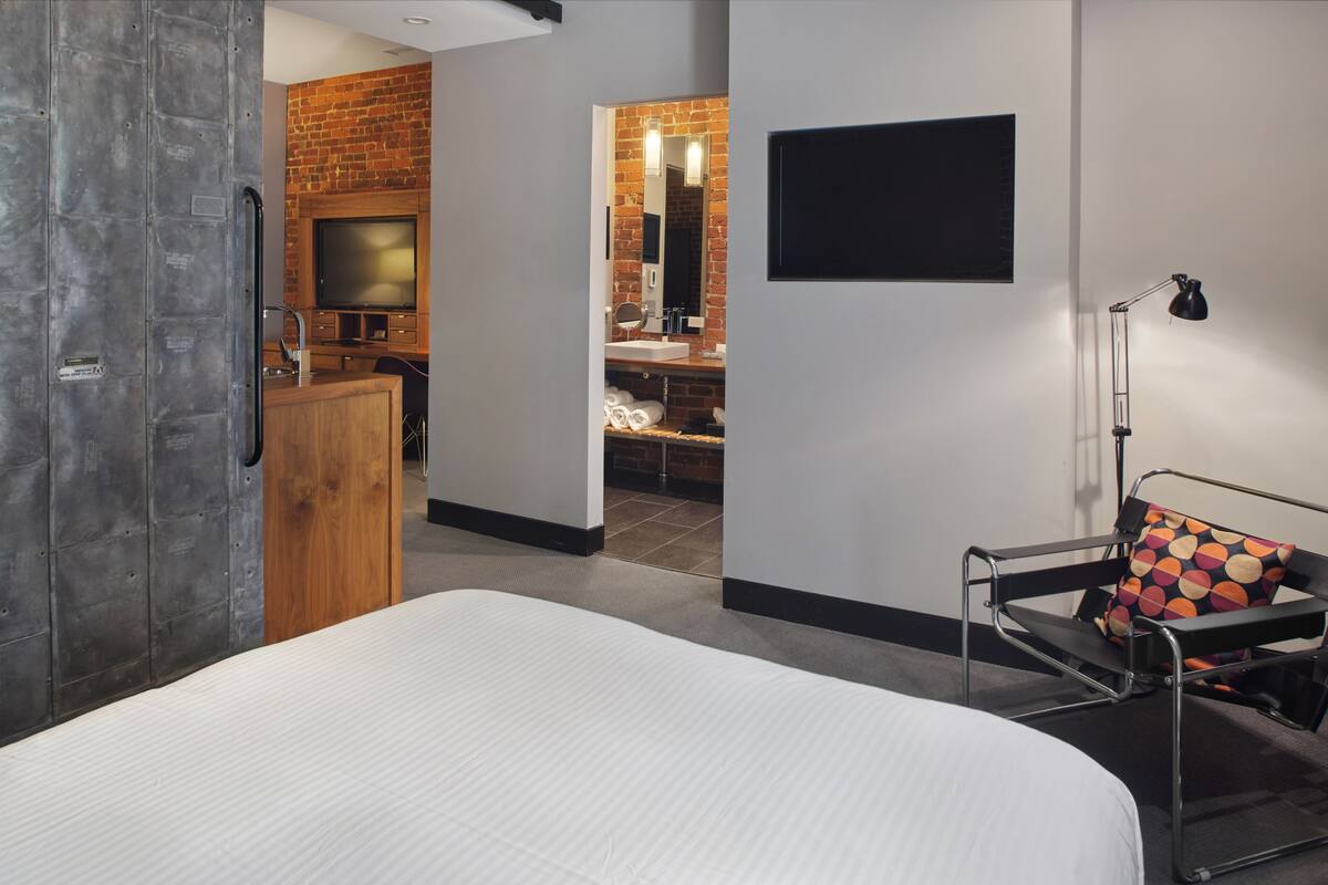 A modern hotel room with a large bed, a wooden wardrobe, a brick accent wall, a TV, and a desk area with a chair and a lamp.