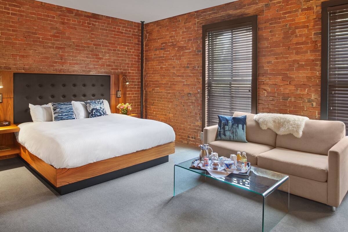 A modern hotel room with a brick wall, a large bed with a wooden frame, a beige sofa, a glass coffee table, and a nightstand with a lamp.