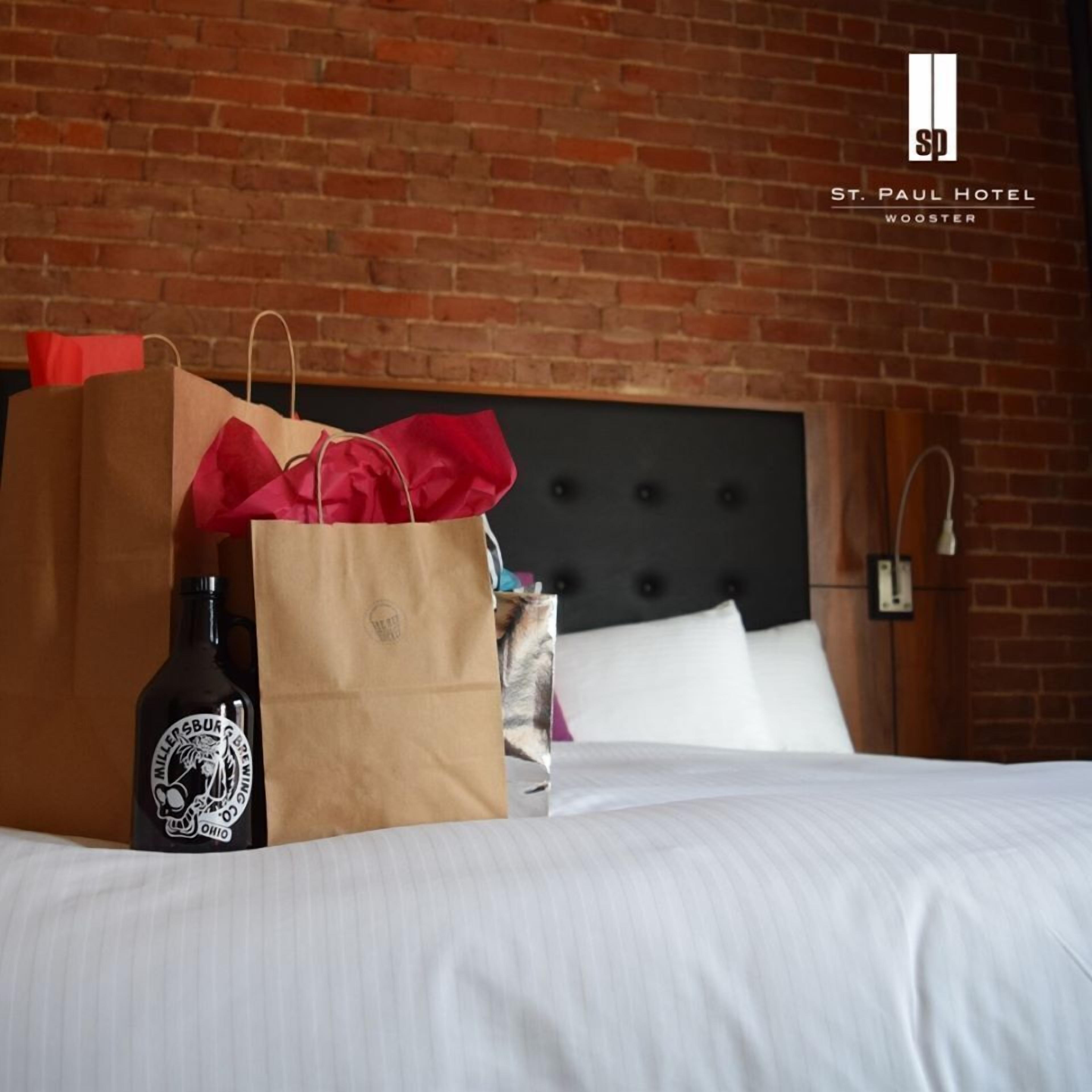 A bed with a headboard, a brown paper bag, a red gift bag, and a bottle of beer in a hotel room with brick walls.