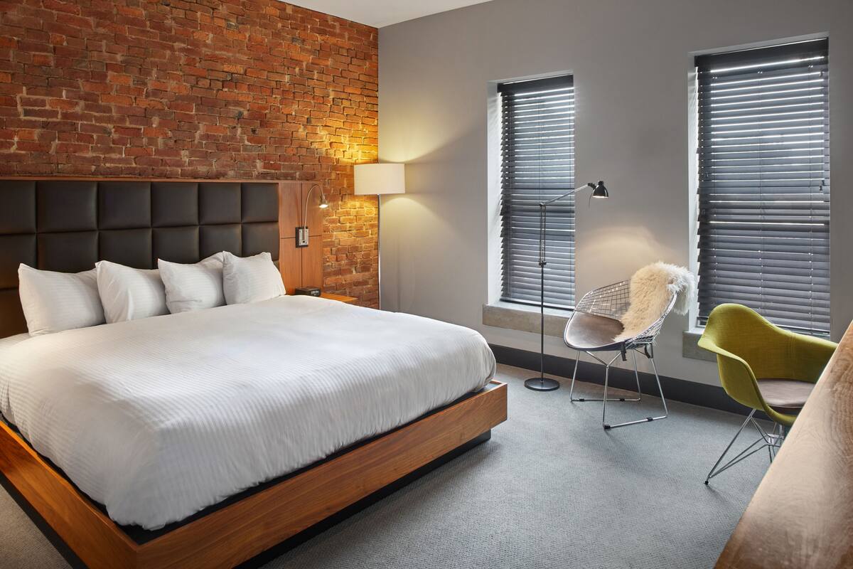 A modern bedroom with a brick wall, a large bed with white bedding, a floor lamp, and a green chair.