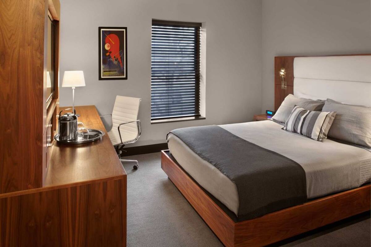 A modern hotel room with a wooden bed frame, a desk with a lamp, a chair, a window with blinds, and a wall-mounted artwork.