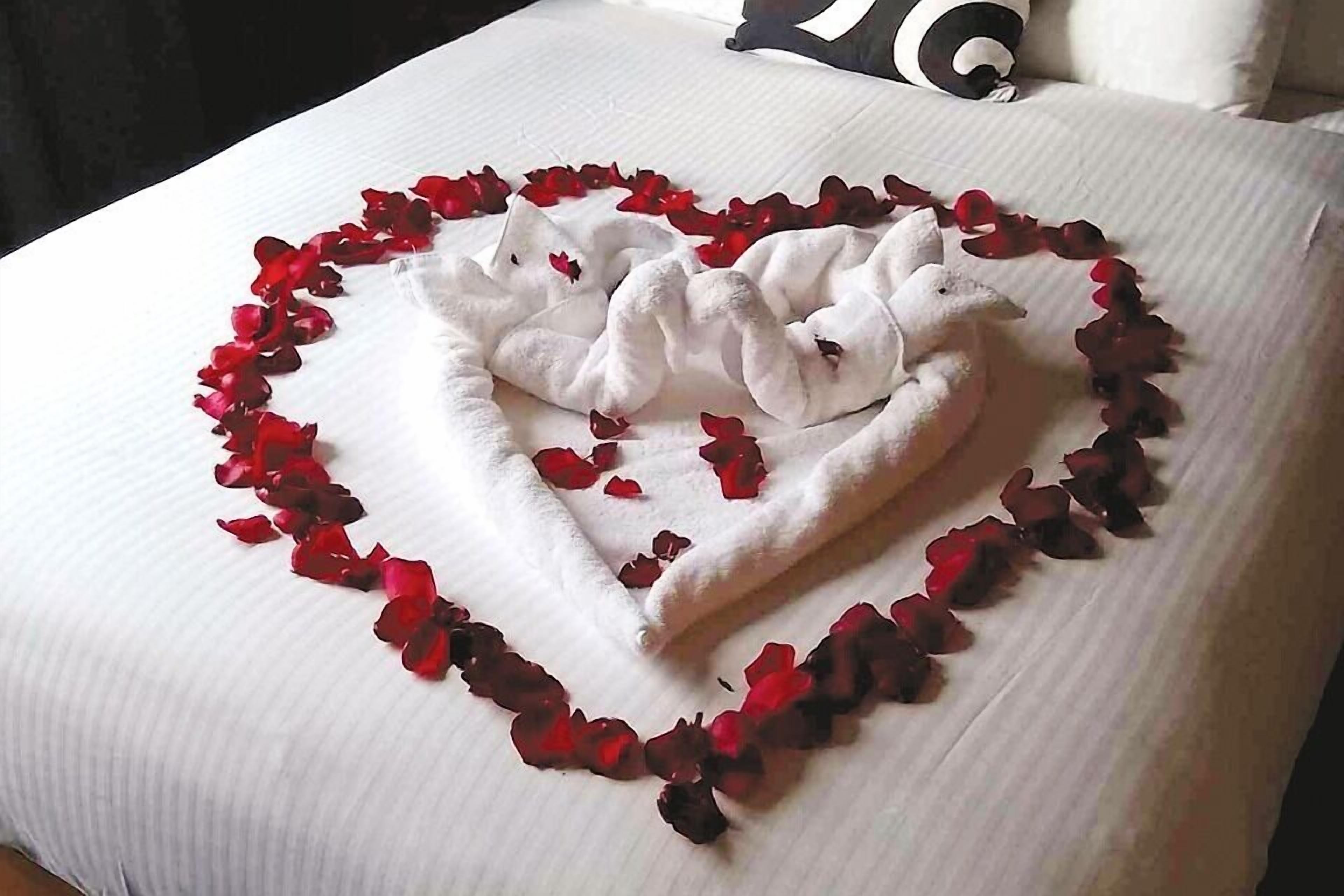 A bed with a heart-shaped towel arrangement and red rose petals.