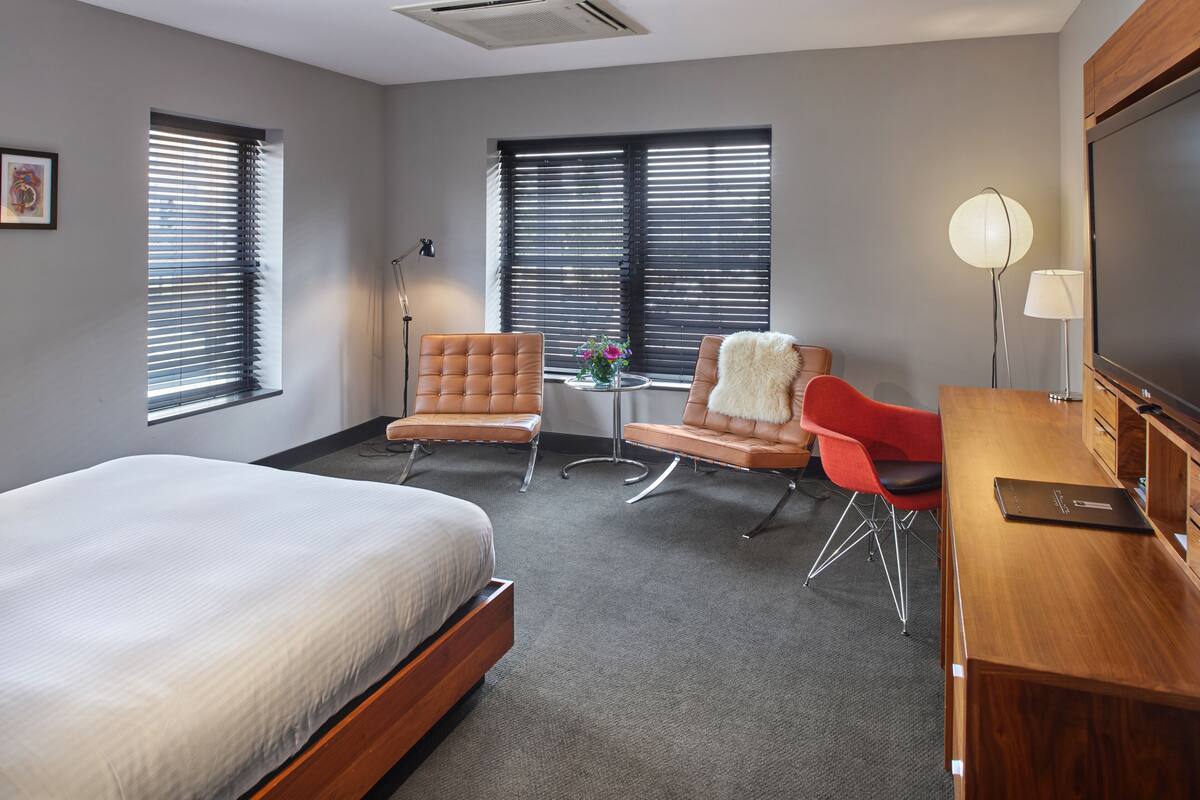 A hotel room with a large bed, two armchairs, a small table with a vase, and a desk with a lamp and a laptop.