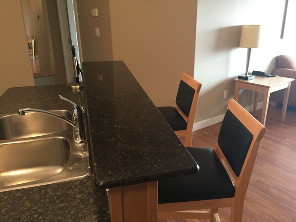 A kitchen with a granite countertop, stainless steel sink, and wooden chairs with black cushions.