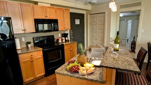 A kitchen with wooden cabinets, a black microwave, a stove, a sink, and a breakfast bar with fruit and a bottle of wine. - Clearwater Residence Hotel Timberlea (Fort McMurray)