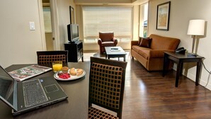 A hotel room with a dining table, a laptop, a plate of food, a TV, a sofa, and a window with blinds. - Clearwater Residence Hotel Timberlea (Fort McMurray)