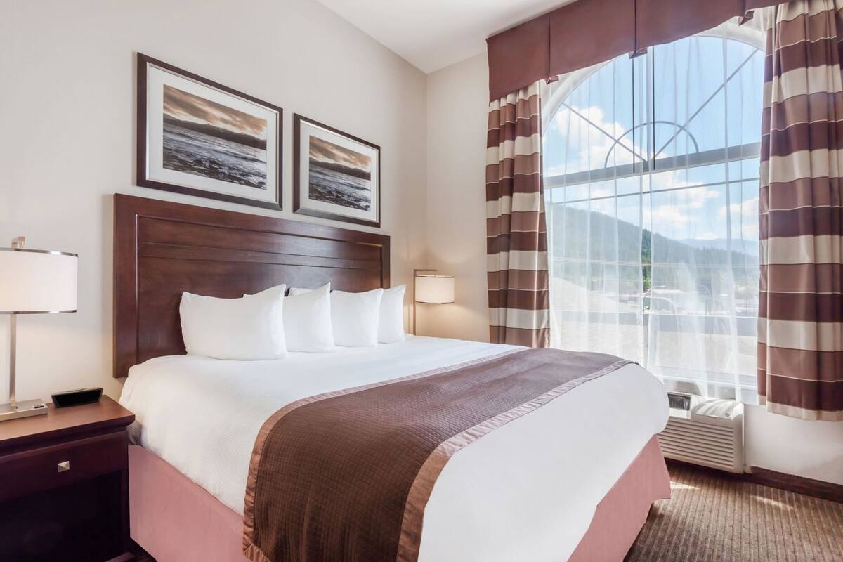 Habitación de hotel con una cama grande, dos cuadros en la pared, una mesita de noche con lámpara y una ventana con cortinas.