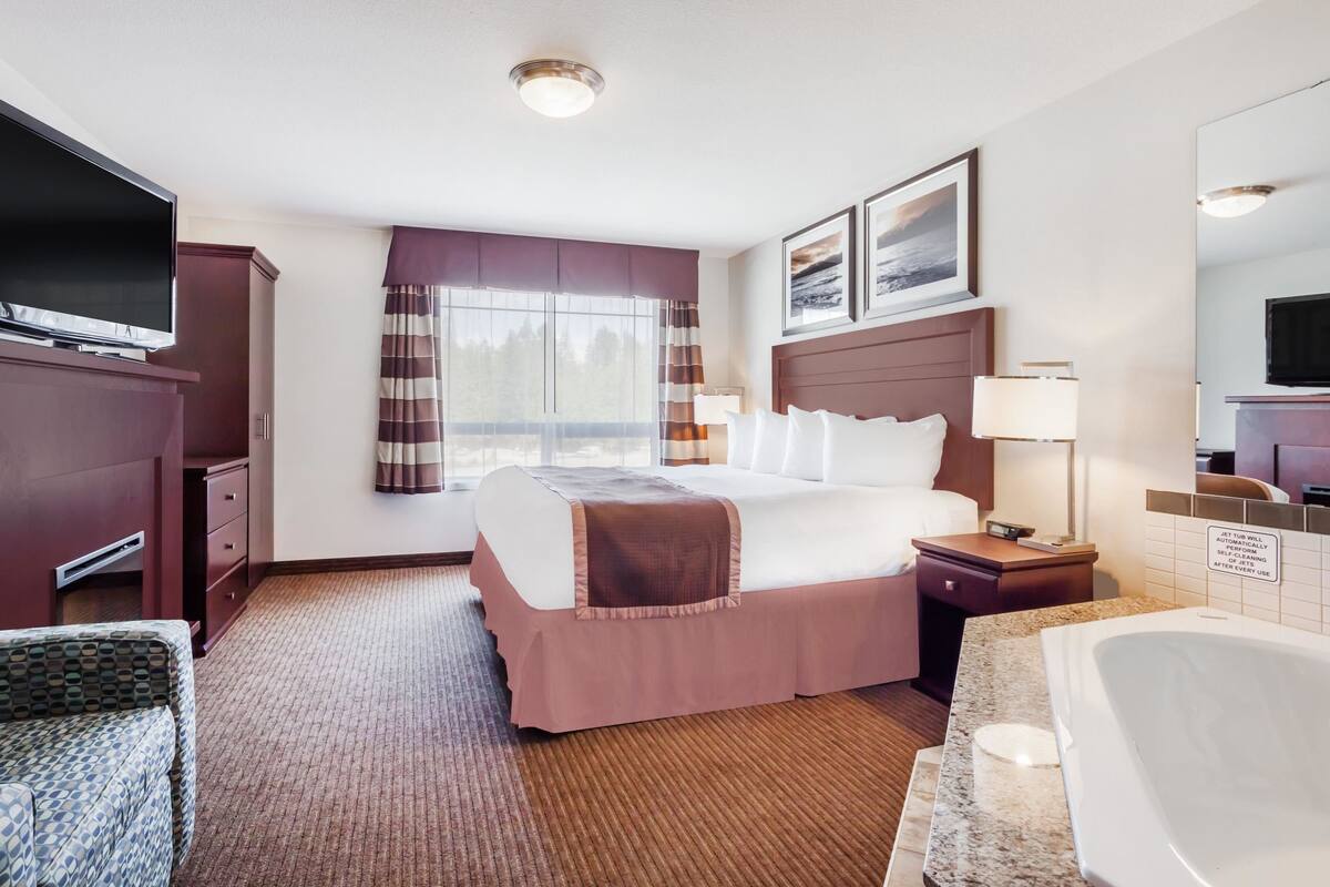 Habitación de hotel con cama, televisor, escritorio, silla, baño con bañera y ventana con cortinas.
