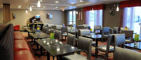 A hotel dining area with multiple tables and chairs, a long red bench, and a clock on the wall.