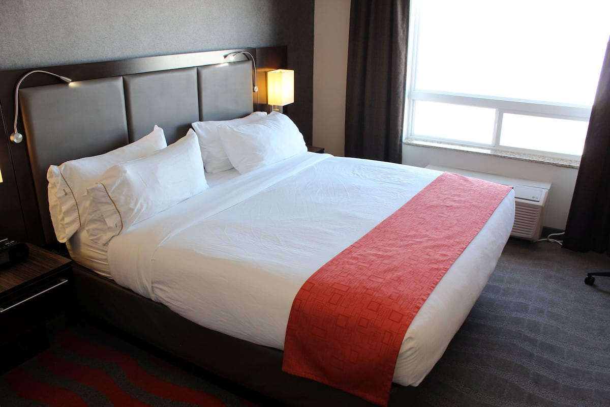 Una cama bien hecha con ropa de cama blanca y roja, un cabecero con una lámpara y una ventana con cortinas.