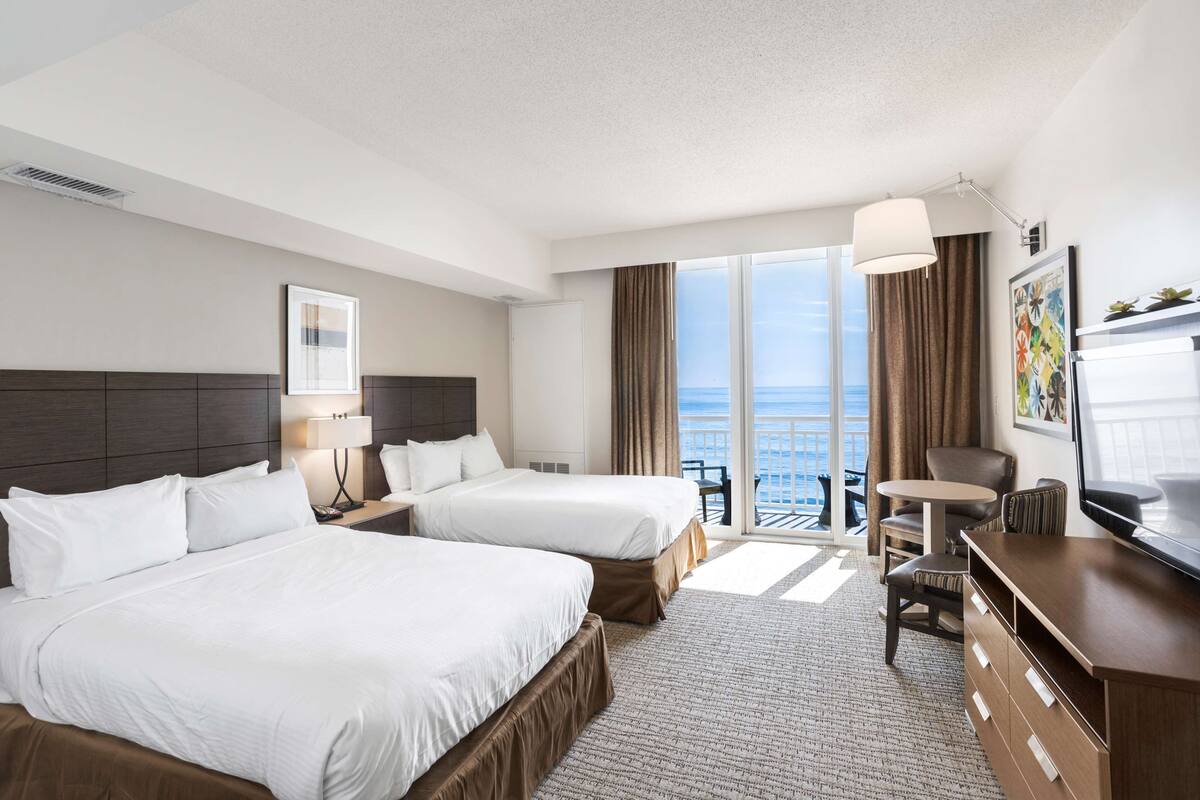 A hotel room with two beds, a desk, a chair, a TV, and a view of the ocean.