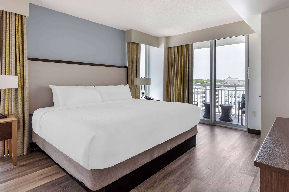 A hotel room with a large bed, a nightstand, a desk, and a view of a cityscape through large windows.