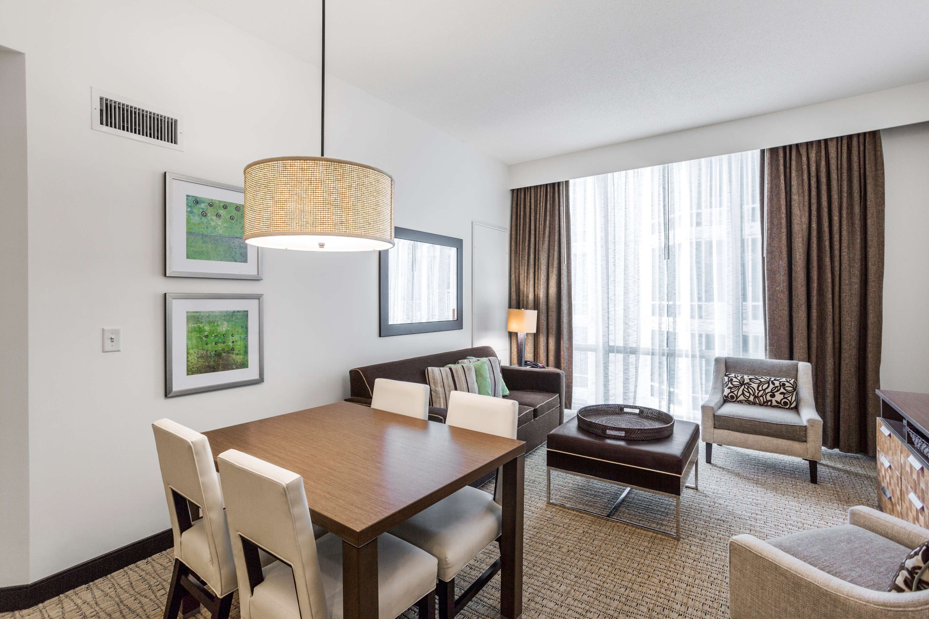 Een hotelkamer met een eettafel, stoelen, een bank, een fauteuil, een lamp en grote ramen voorzien van gordijnen.