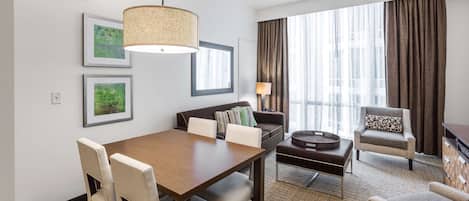 Een hotelkamer met een eettafel, stoelen, een bank, een fauteuil, een lamp en grote ramen voorzien van gordijnen.