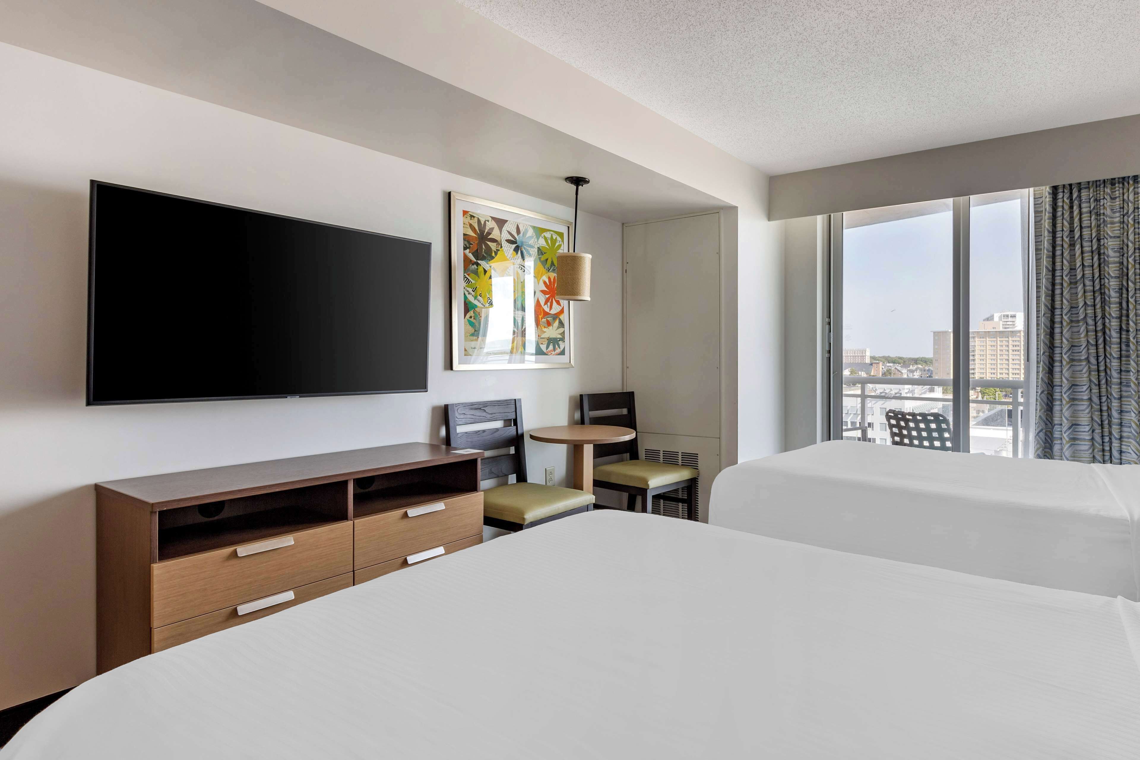 A hotel room with two beds, a flat-screen TV, a small table with chairs, and a large window with a city view.