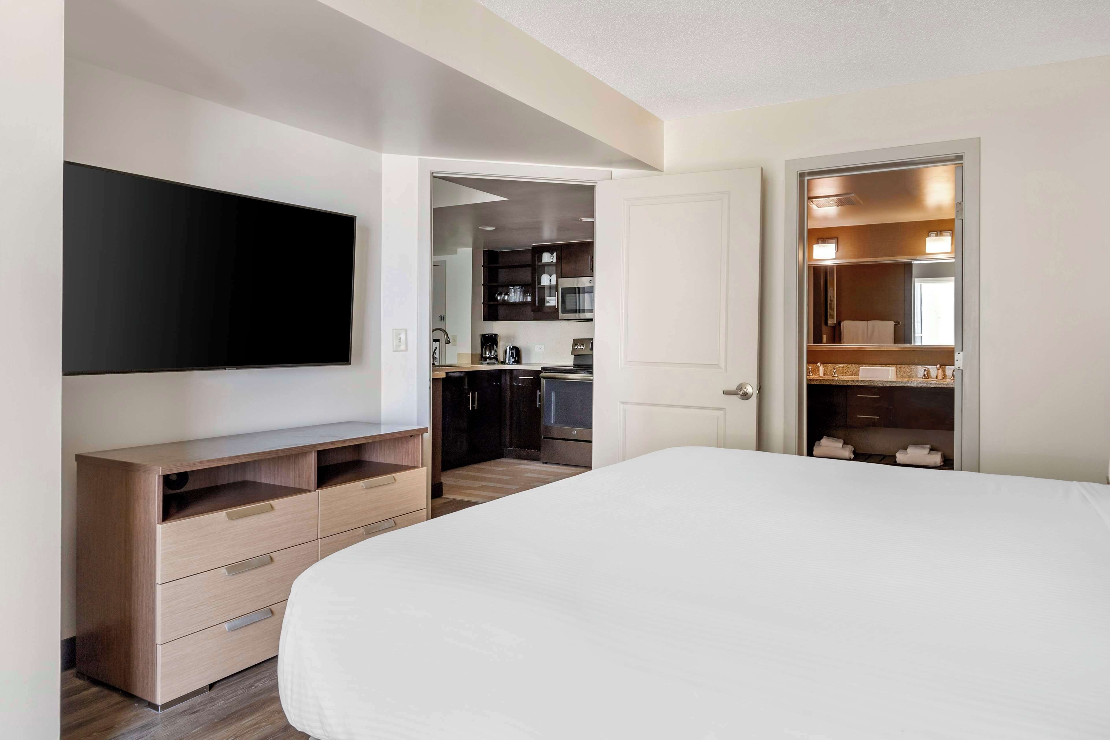 A bedroom with a large bed, a TV mounted on the wall, a wooden dresser, and a kitchen area visible through an open door.