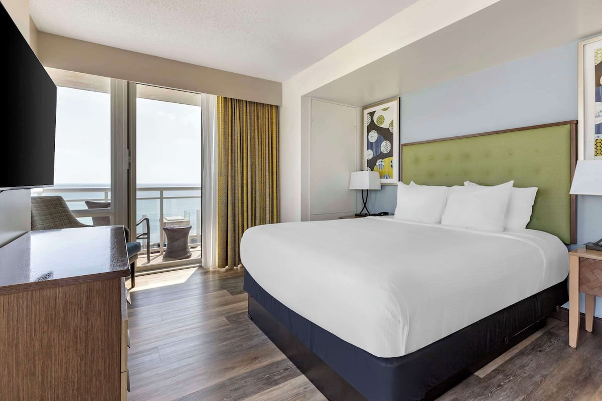 A hotel room with a large bed, a desk, a chair, a balcony with curtains, and a view of the sea.