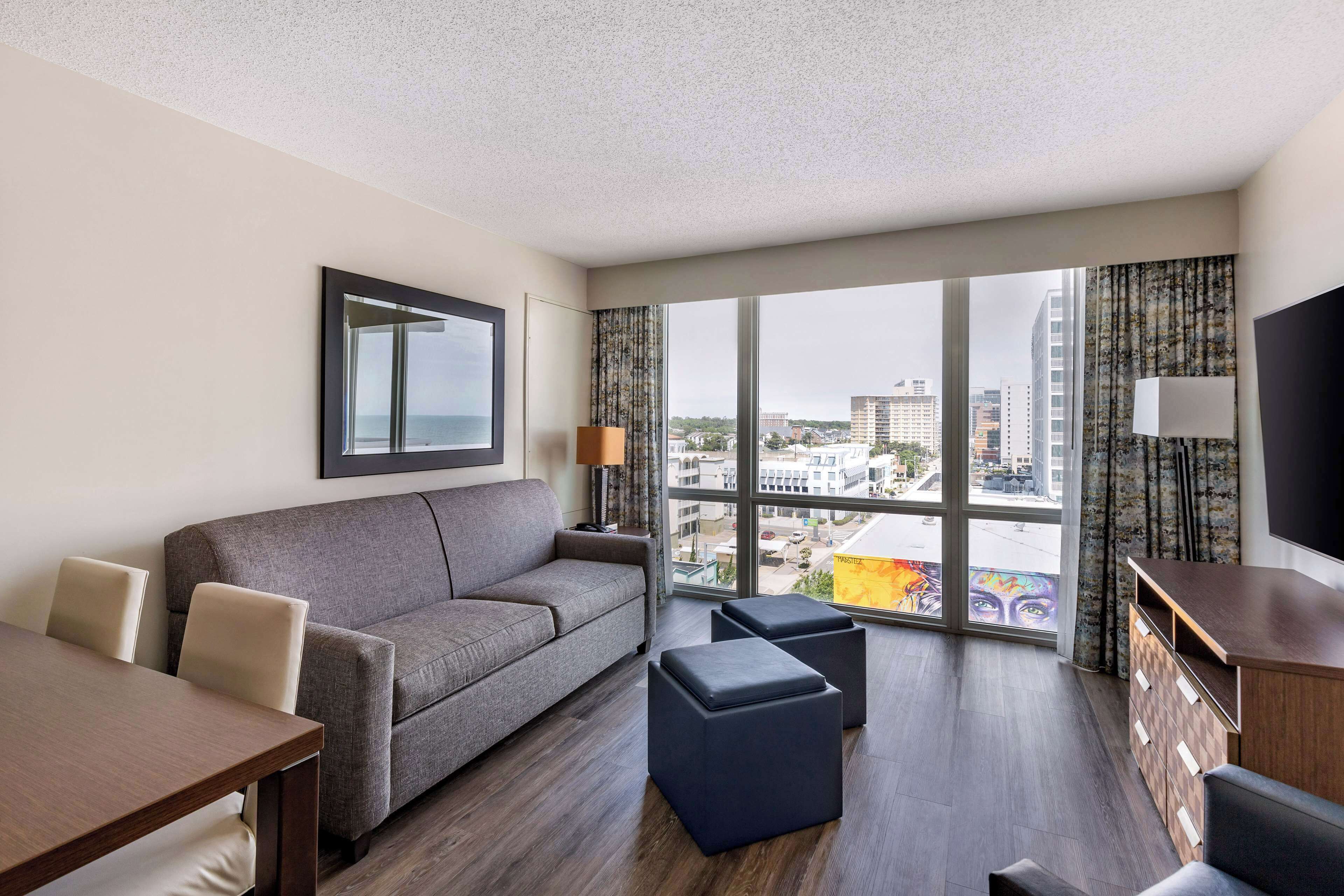 A modern hotel room with a grey sofa, a wooden dining table, and a large window offering a city view.
