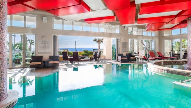 Zona de piscina con agua azul cristalina, vigas de techo rojas y mobiliario exterior.