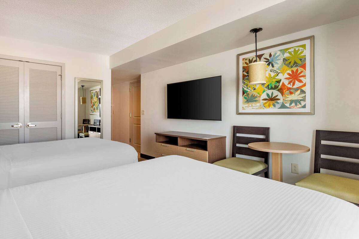 A hotel room with two beds, a TV, a small table, and a colorful artwork on the wall.