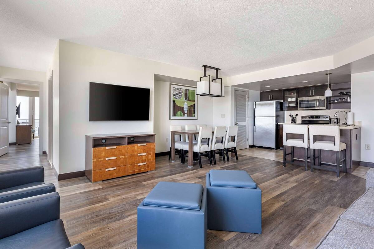 A modern living room with a flat-screen TV, a dining area with a table and chairs, a kitchen with appliances, and a seating area with ottomans.