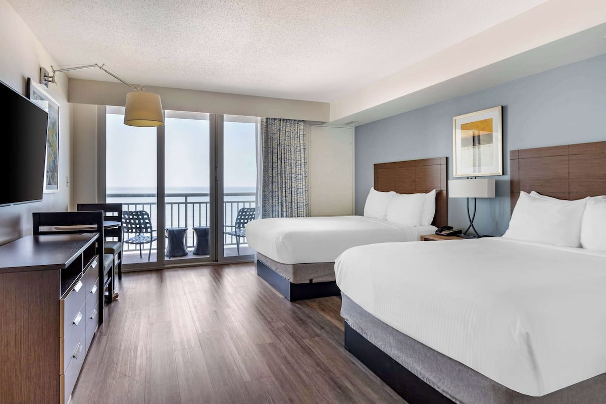 A hotel room with two beds, a desk, a TV, and a balcony with a view.