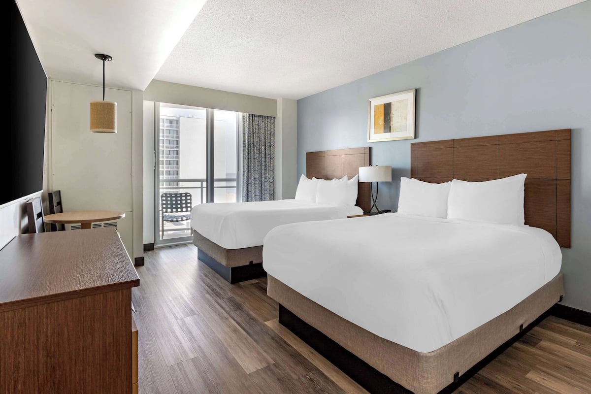 A hotel room with two beds, a desk, a chair, a lamp, and a window with a view of the city.