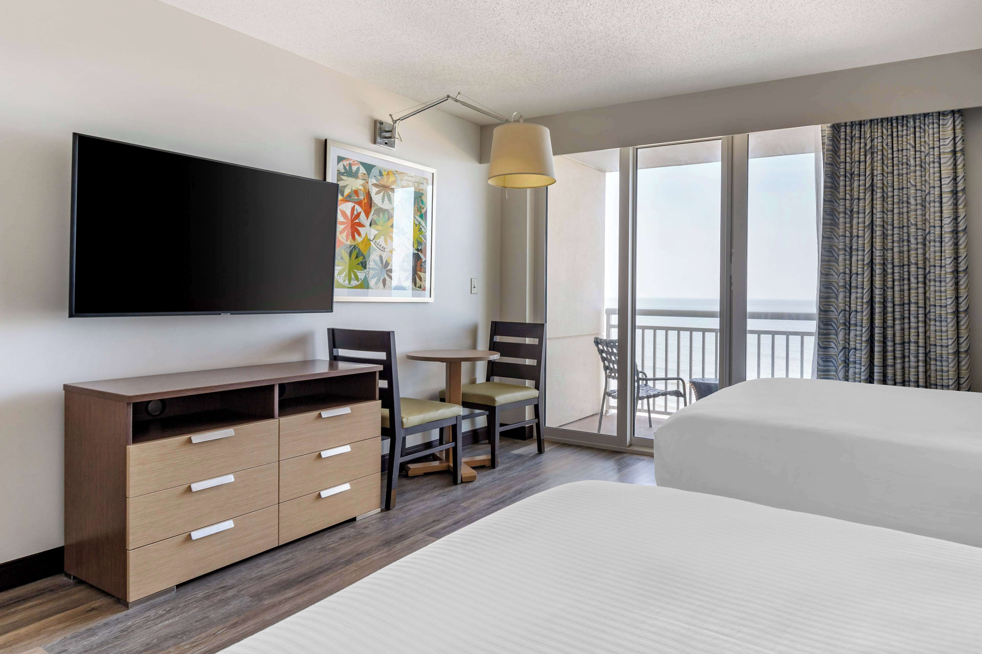 A hotel room with a large bed, a flat-screen TV, a desk with chairs, and a balcony with a view.