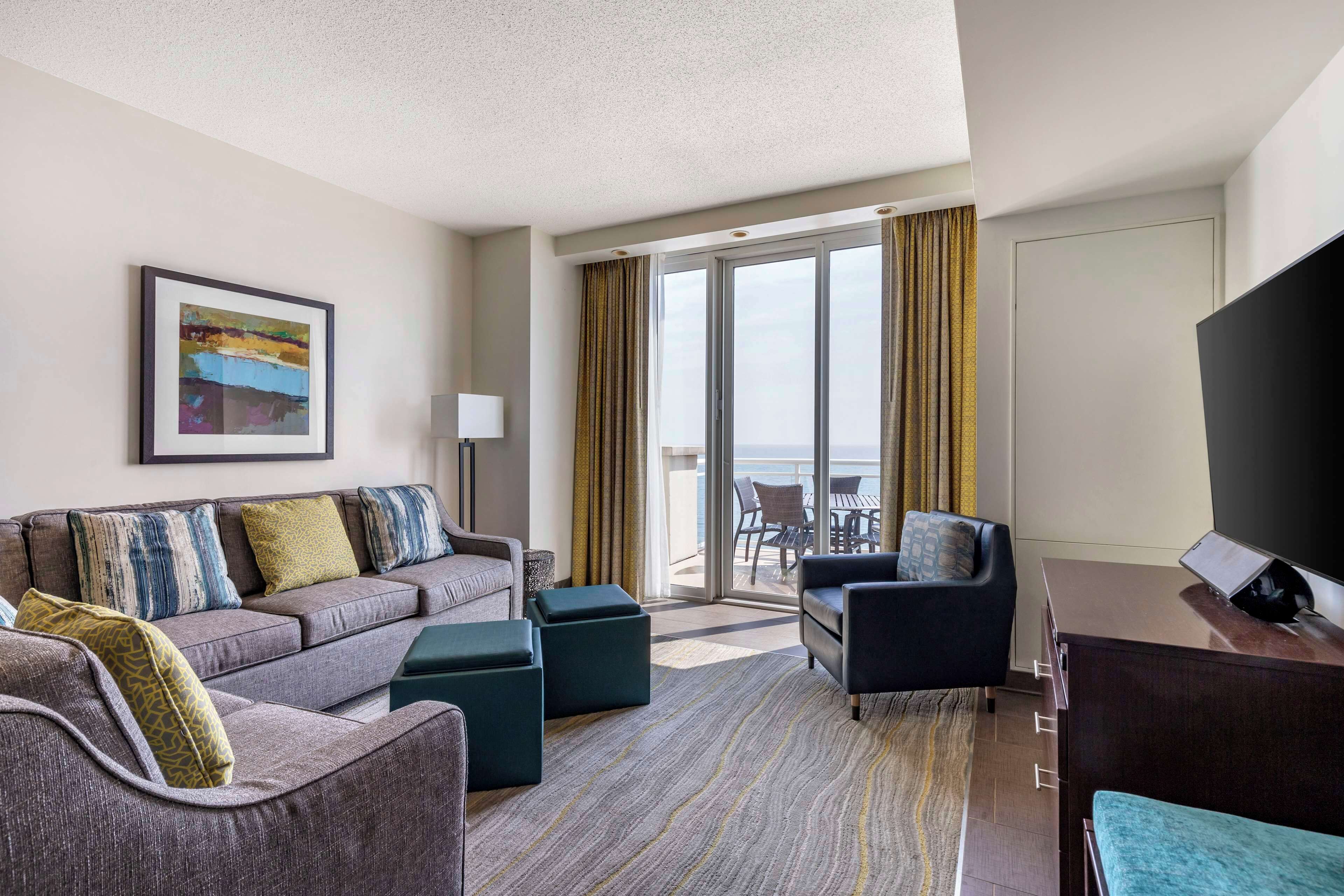 Ein Hotelzimmer mit grauem Sofa, einem schwarzen Sessel, einem Fernseher und einem Balkon mit Blick.