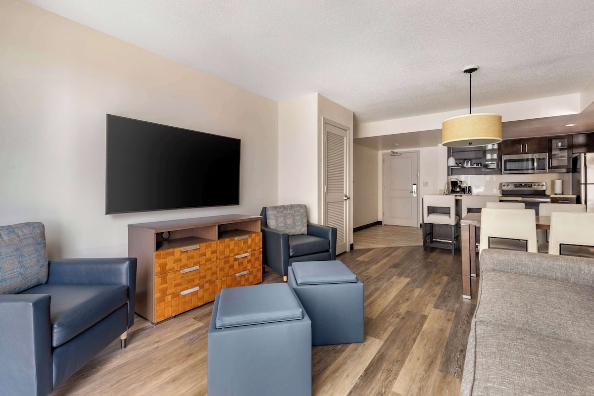 A modern living room with a flat-screen TV, sofas, ottomans, a wooden cabinet, and a dining area with a kitchen in the background.