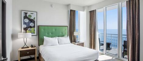 A hotel room with a large bed, a green headboard, a nightstand with a lamp, a framed picture on the wall, and a balcony with a view of the sea.